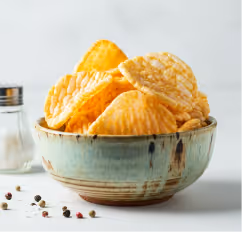 Ceramic bowl filled with ridged potato chips, with a salt shaker and scattered peppercorns nearby on a white surface.