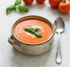 Bowl of creamy tomato soup garnished with basil leaves, with fresh tomatoes and basil in the background and a spoon beside the bowl.