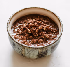 Ceramic bowl filled with creamy chocolate oatmeal topped with crumbled chocolate granola on a white surface.