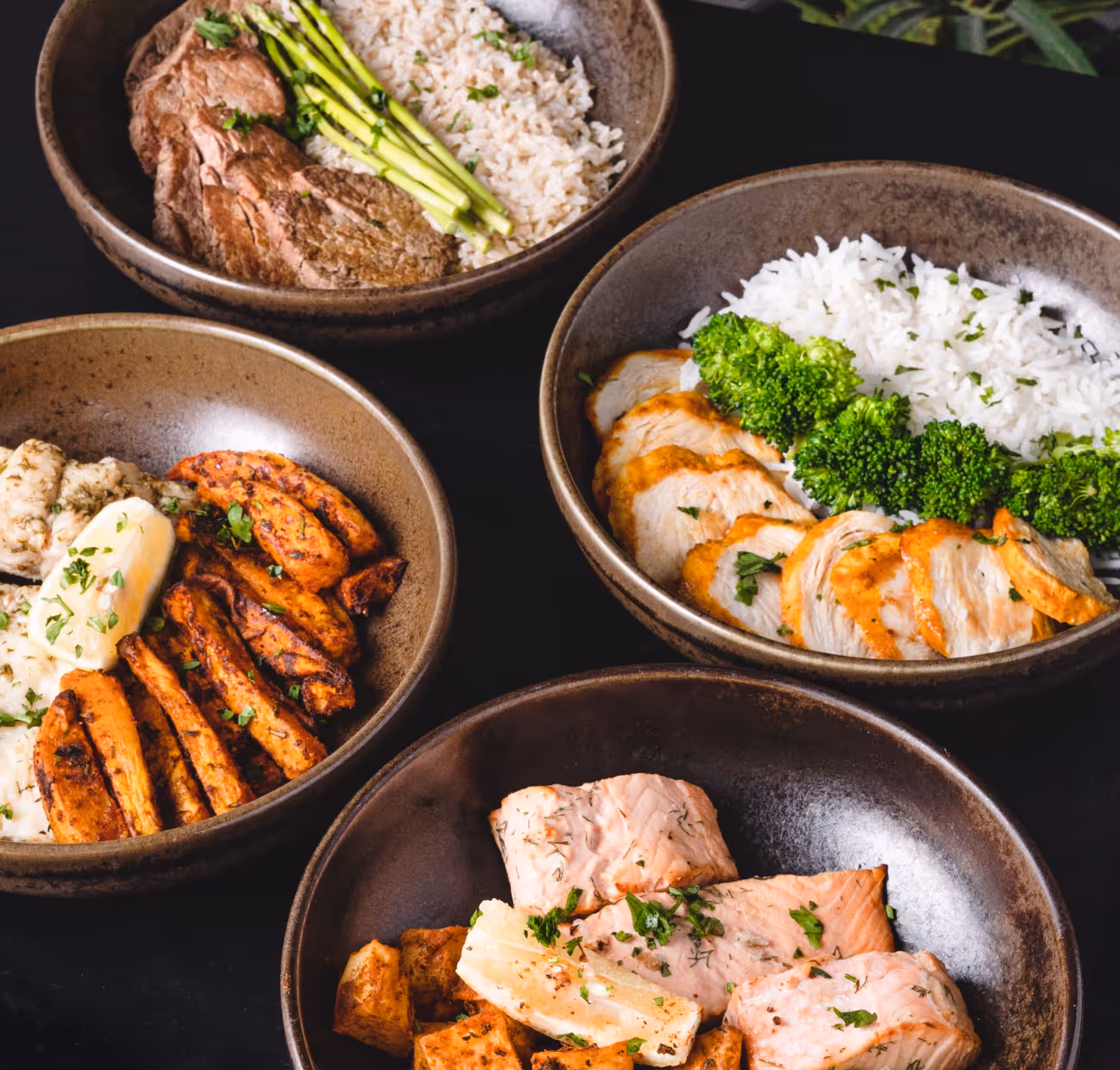 Four bowls containing various meals: steak with rice and asparagus, grilled chicken with rice and broccoli, roasted sweet potatoes with cauliflower, and salmon with roasted vegetables.