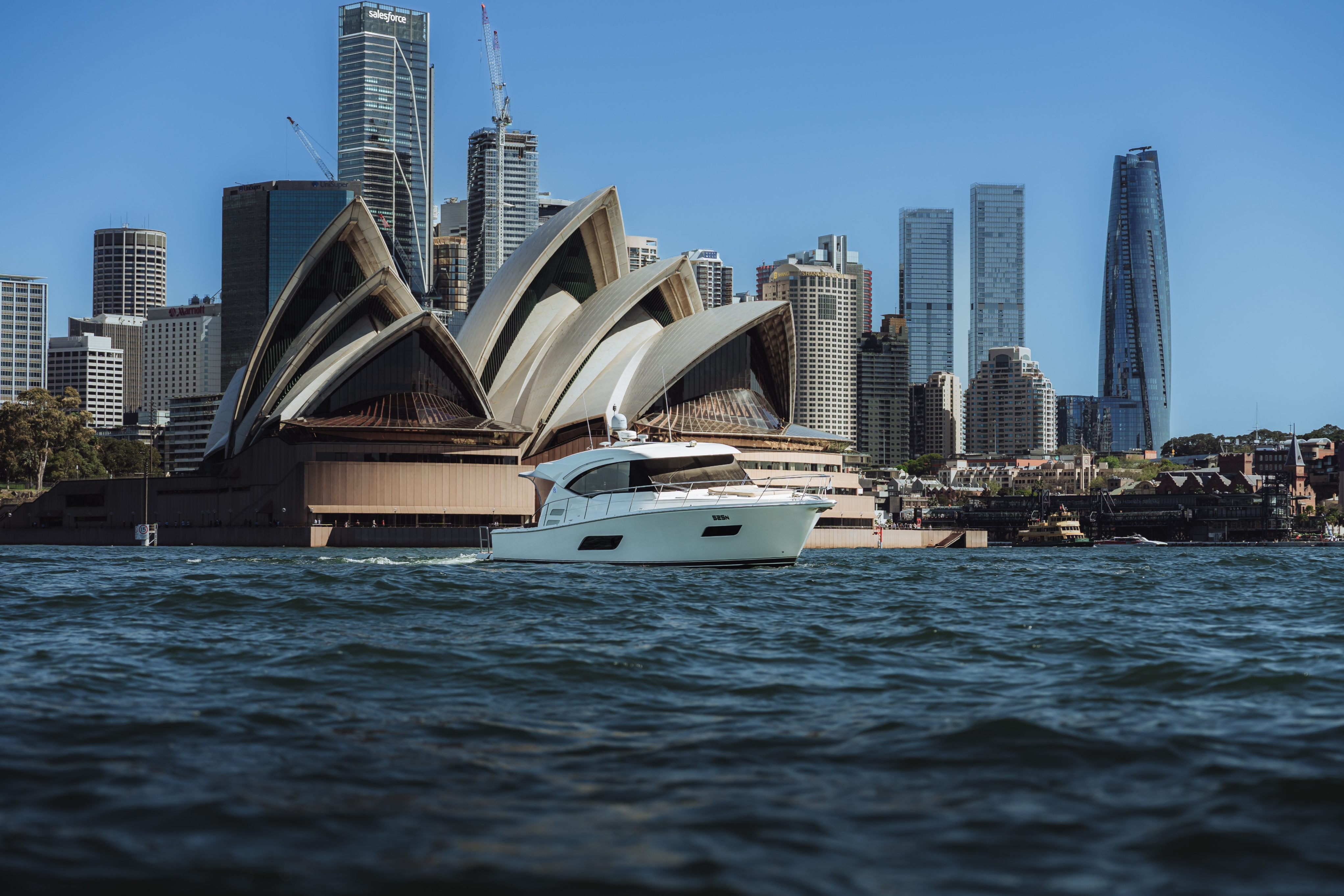 White yacht sailing in front of Sydney Opera House with city skyscrapers and clear blue sky in the background.