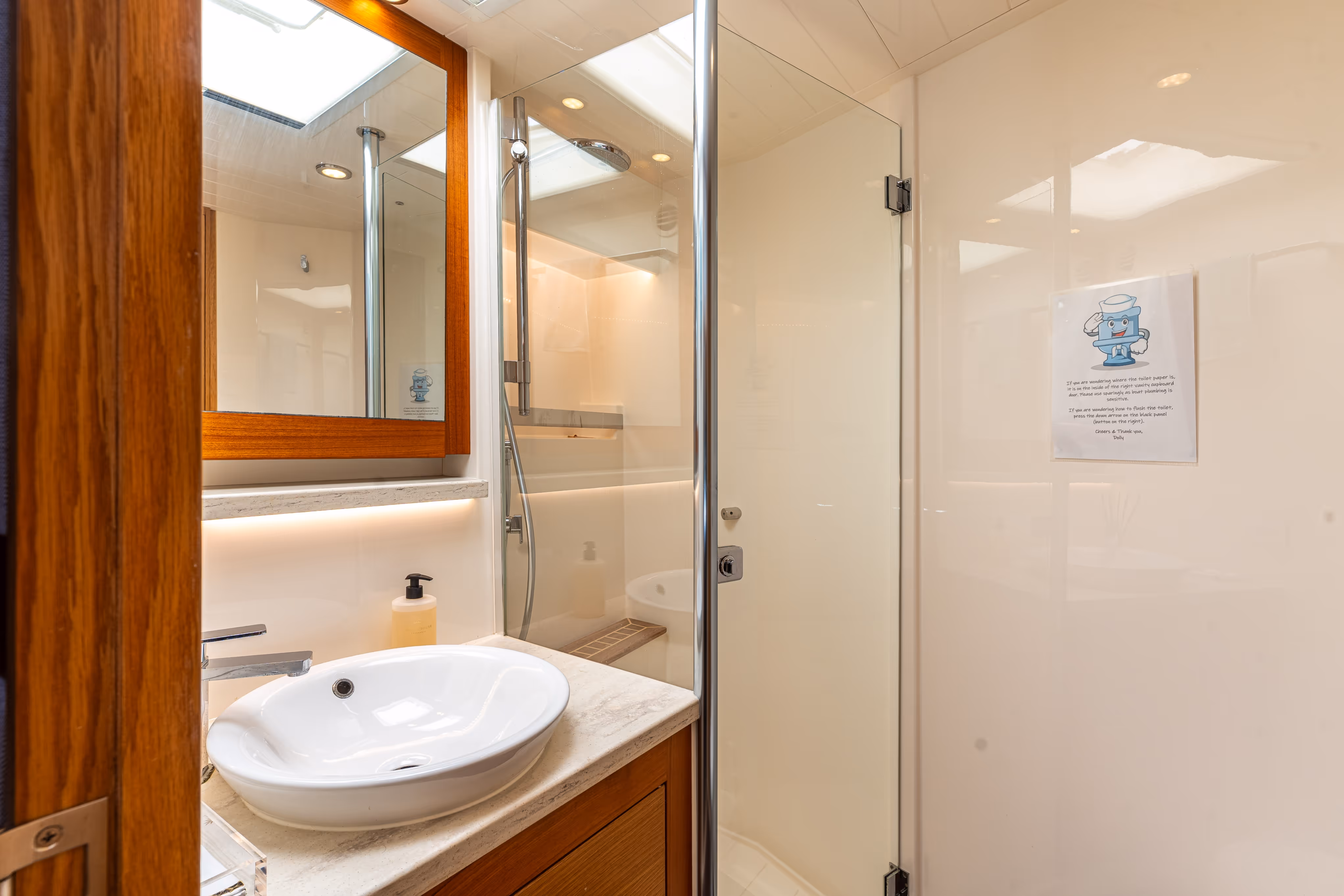 Modern bathroom with a white vessel sink on a marble countertop, wooden framed mirror, glass shower door, and a soap dispenser.