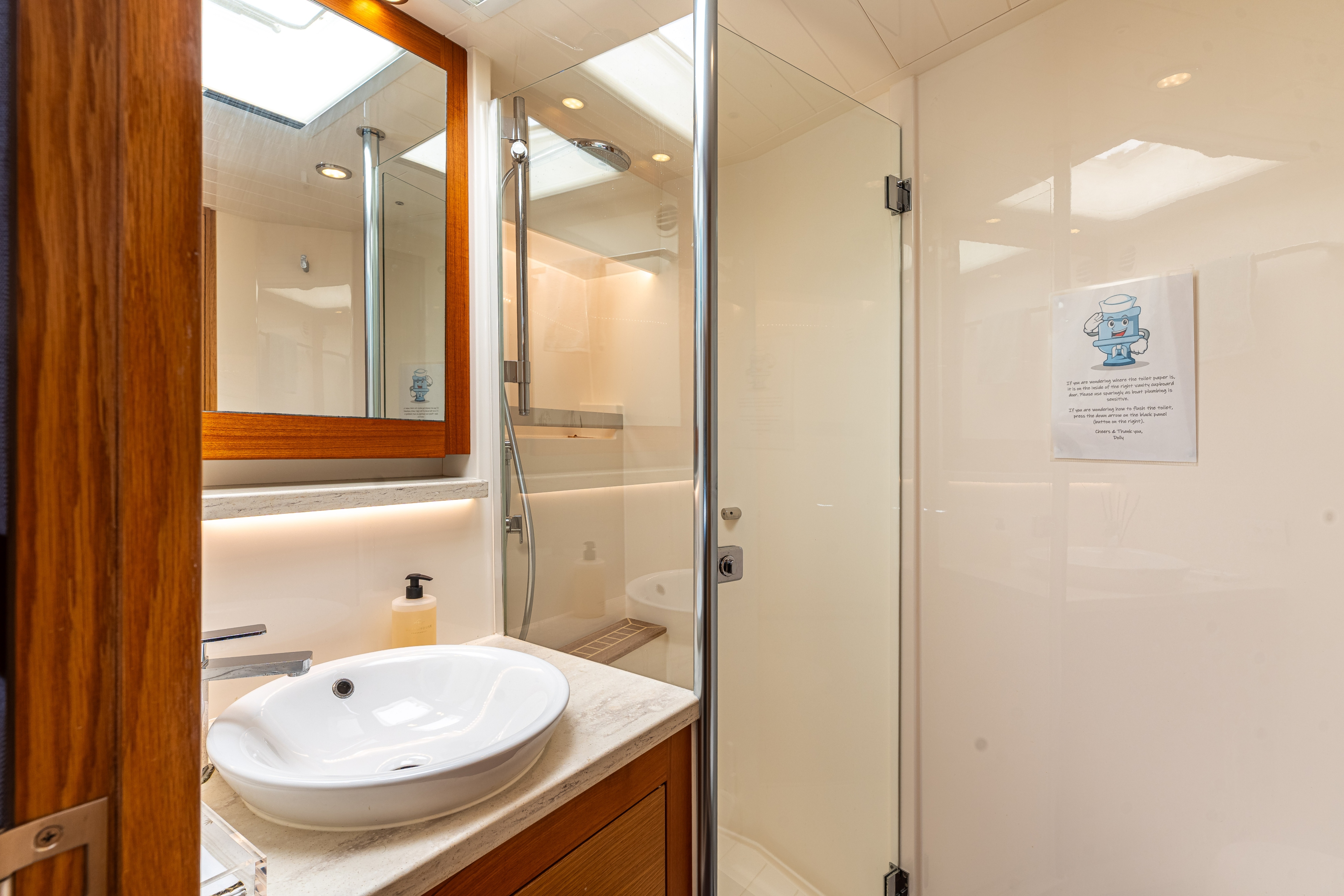 Modern bathroom with a white vessel sink on a marble countertop, wooden framed mirror, glass shower door, and a soap dispenser.