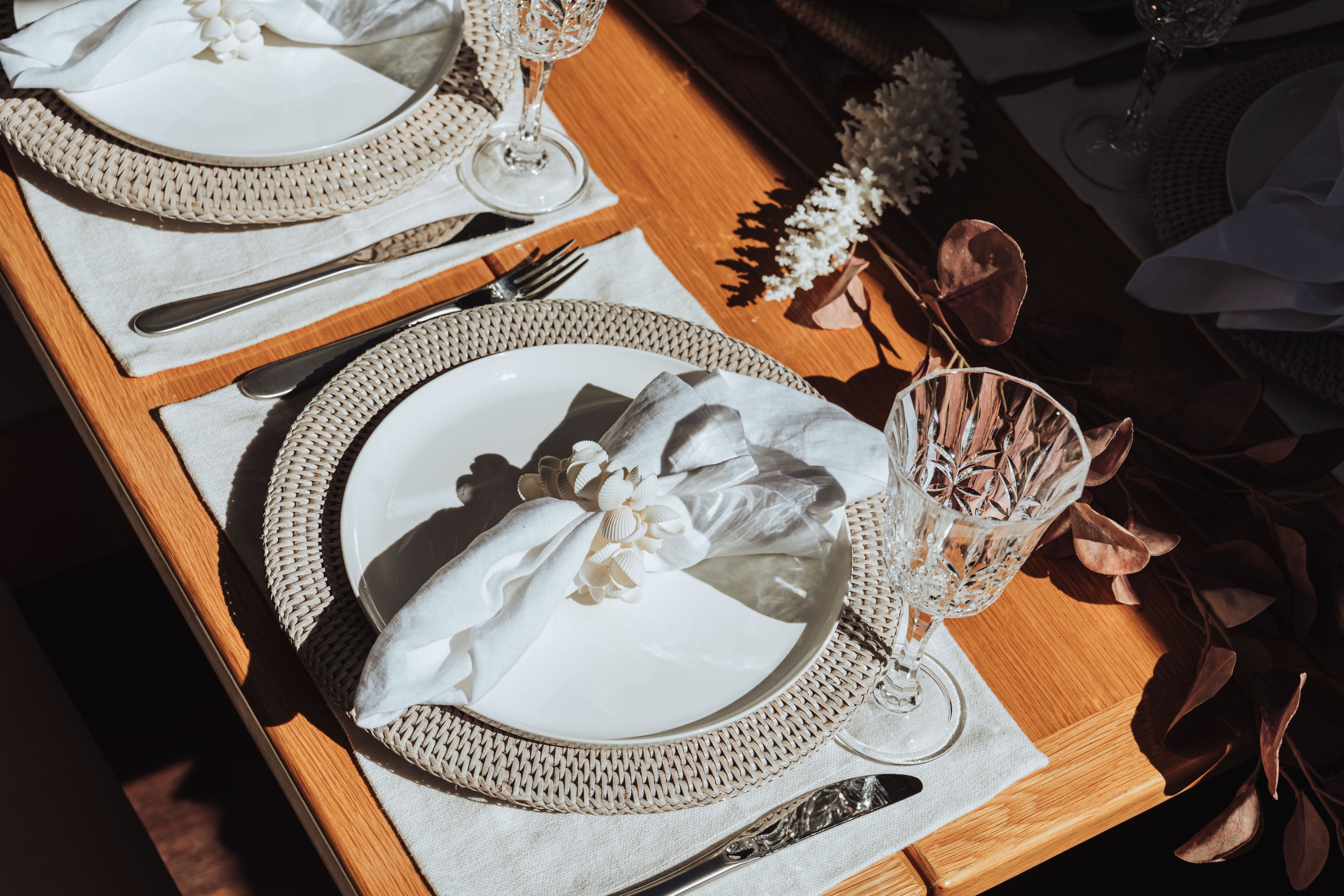 Elegant table setting with white ceramic plates on woven placemats, white linen napkins with decorative rings, crystal wine glasses, and silver cutlery on a wooden table.