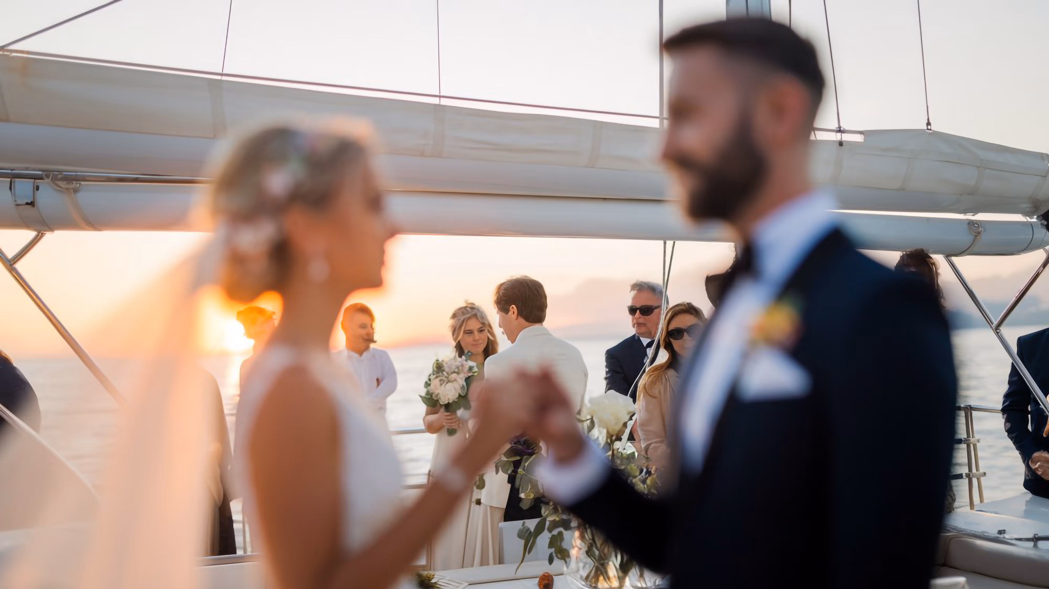 Bride and groom holding hands on a boat at sunset with wedding guests in the background.