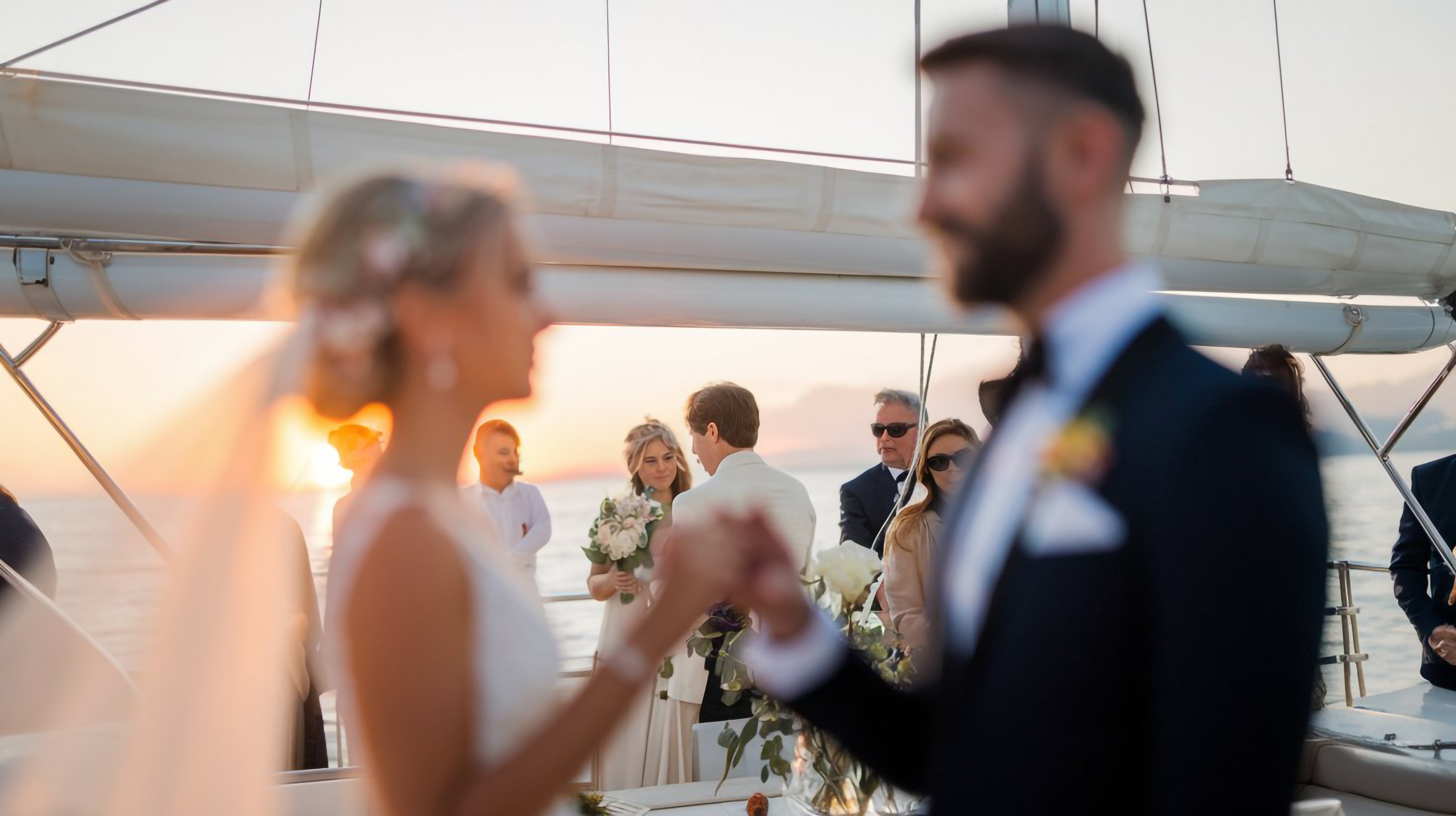 Bride and groom holding hands on a boat at sunset with wedding guests in the background.
