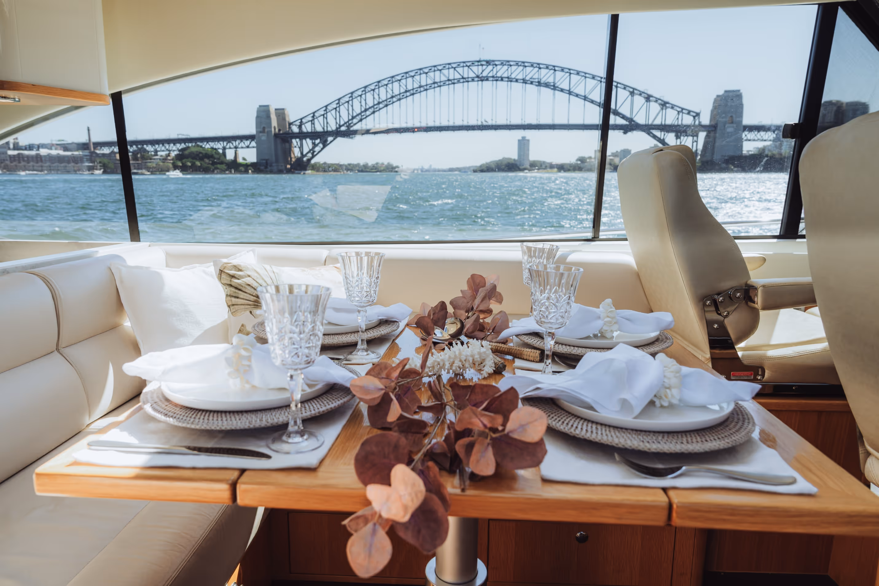 Table set for four with white plates, crystal glasses, and napkins inside a yacht with Sydney Harbour Bridge visible through the window.