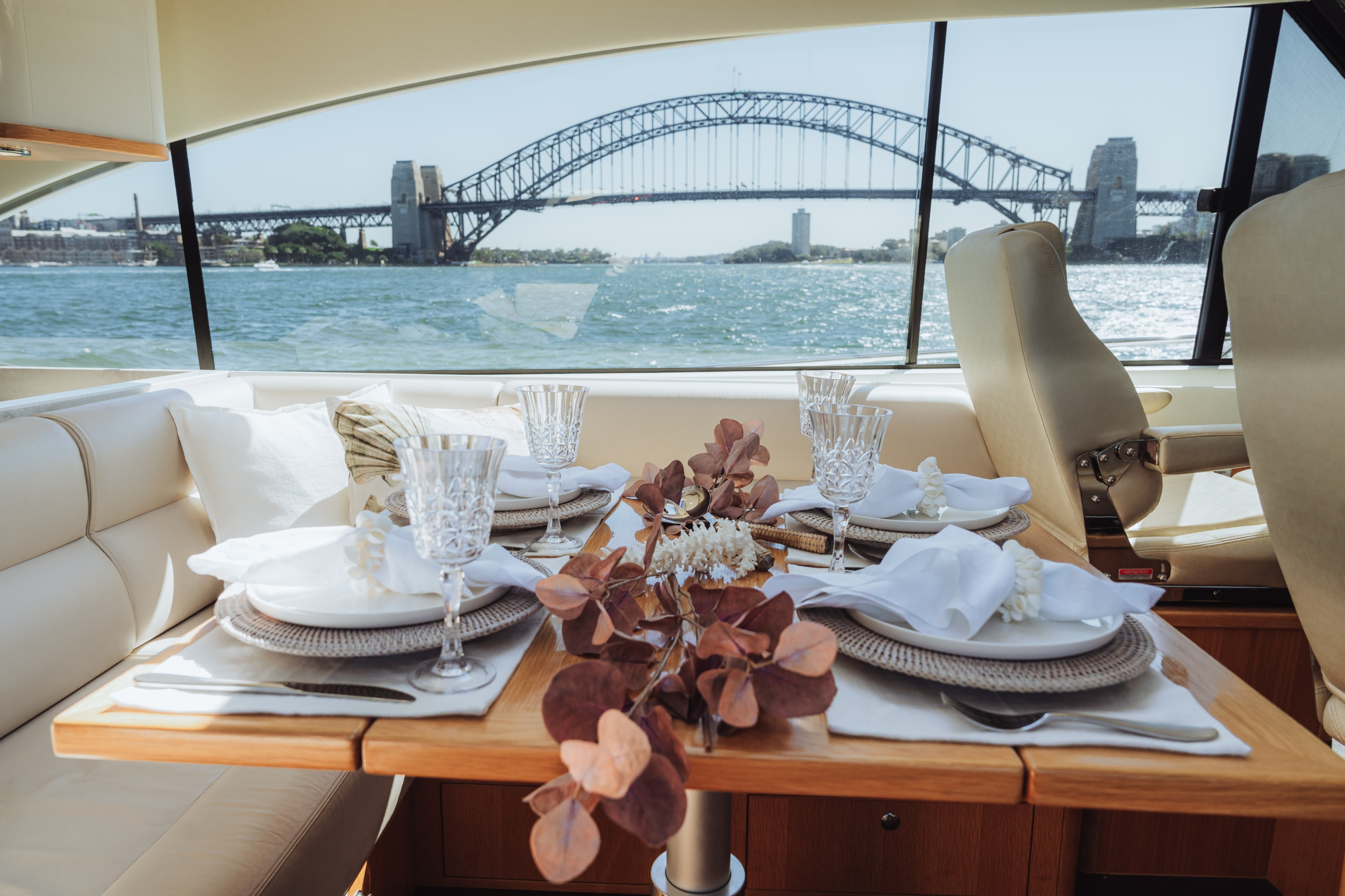 Table set for four with white plates, crystal glasses, and napkins inside a yacht with Sydney Harbour Bridge visible through the window.
