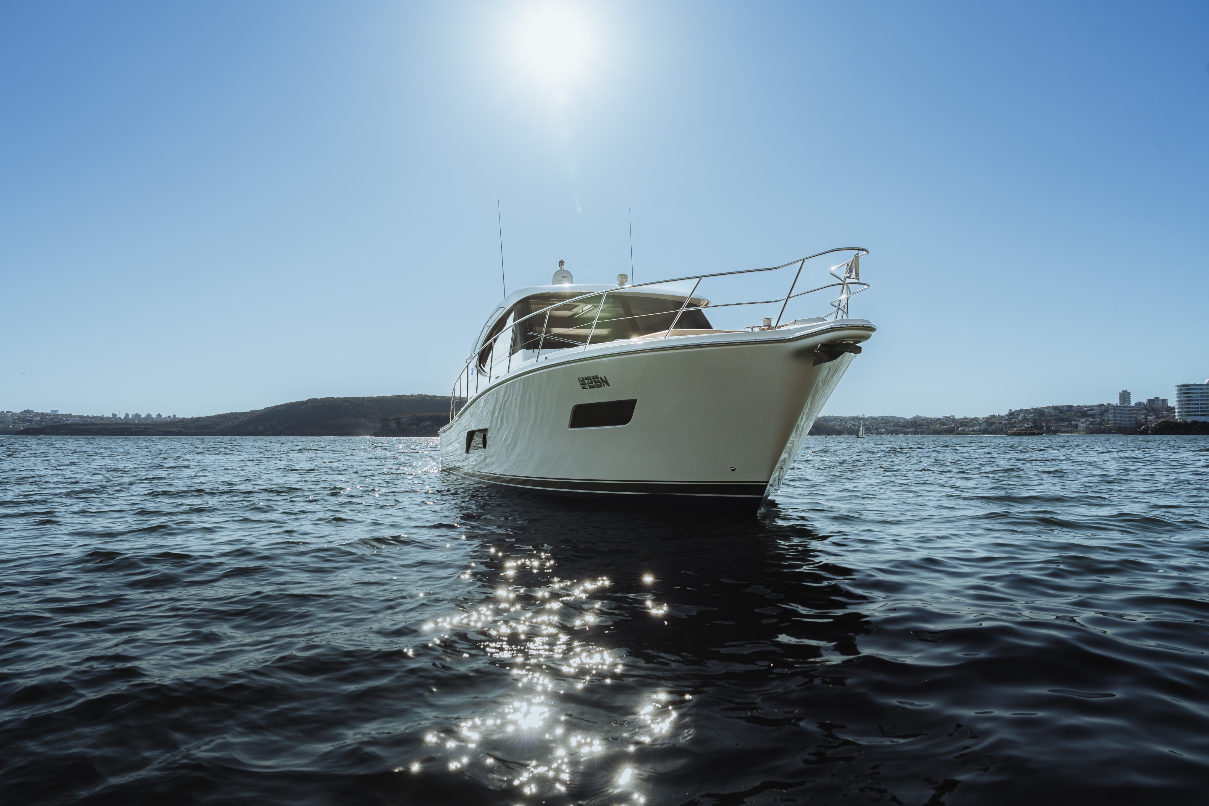 White yacht floating on calm water under a clear blue sky with distant hills and buildings.