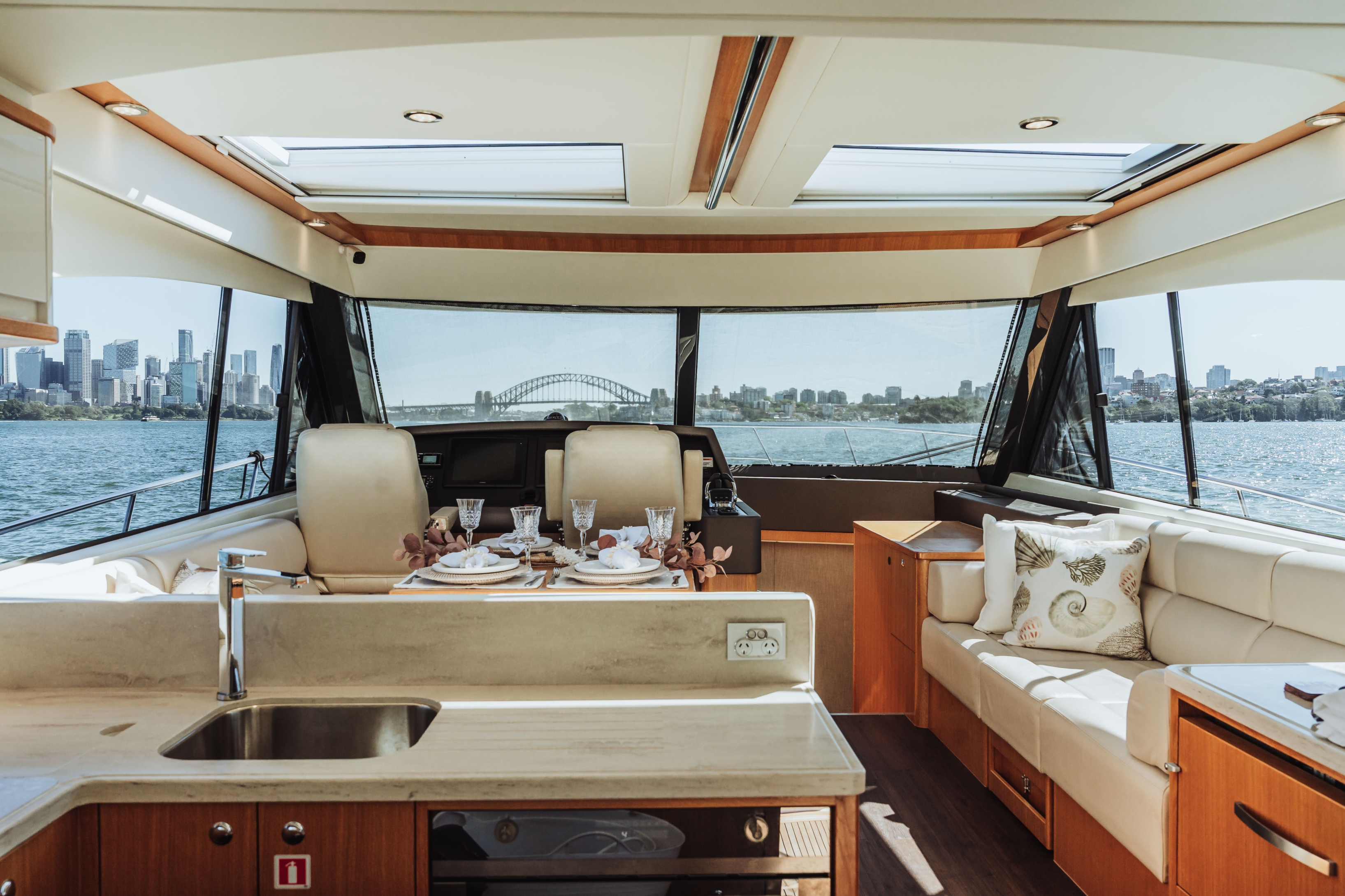 Luxury yacht interior with dining table set for four, cream leather seating, and large windows showing Sydney Harbour Bridge and city skyline.