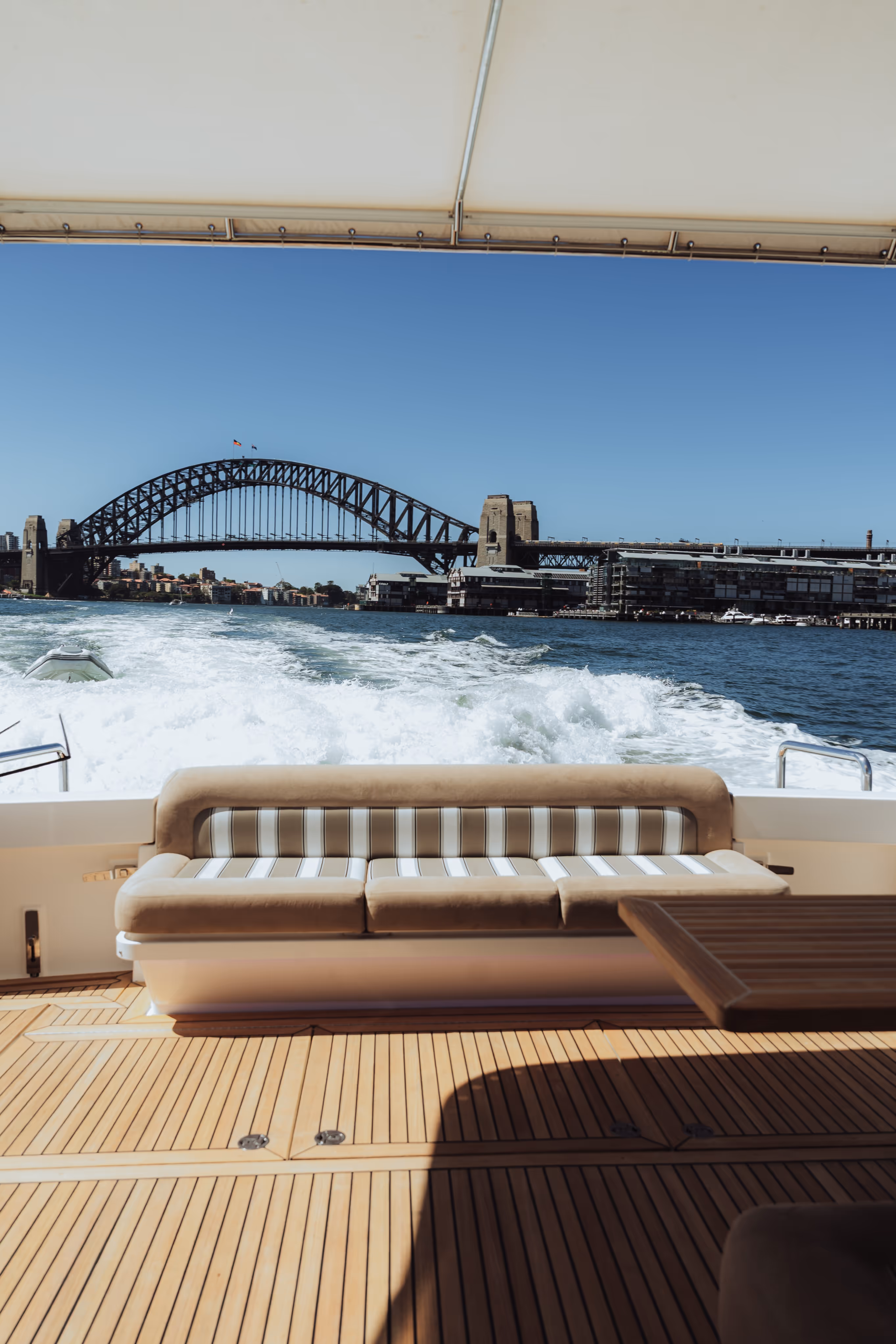 View from a boat showing a cushioned bench and wooden deck with Sydney Harbour Bridge in the background under a clear blue sky.
