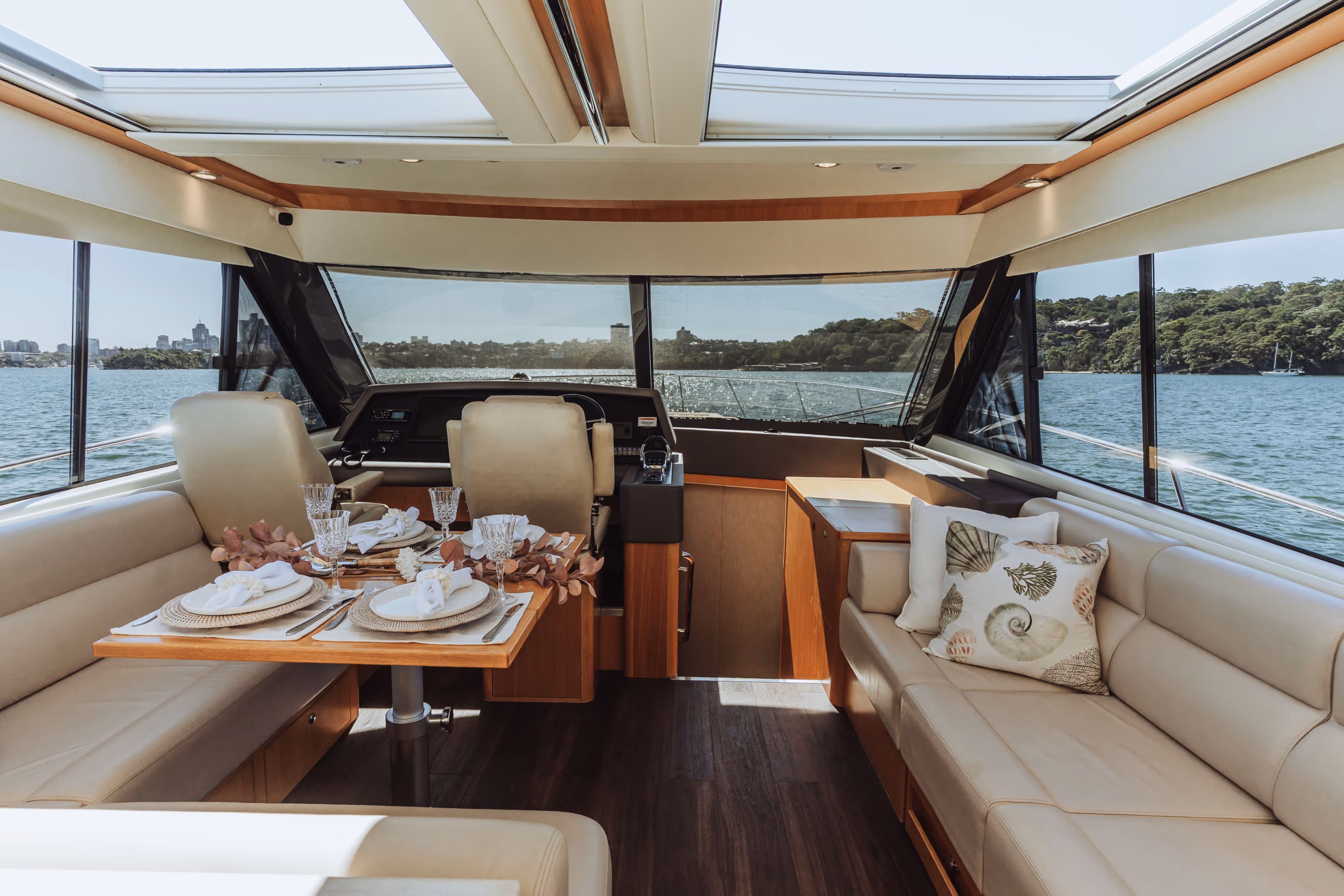Interior of a yacht with a dining table set for four, beige leather seating, and large windows showing water and distant shore.