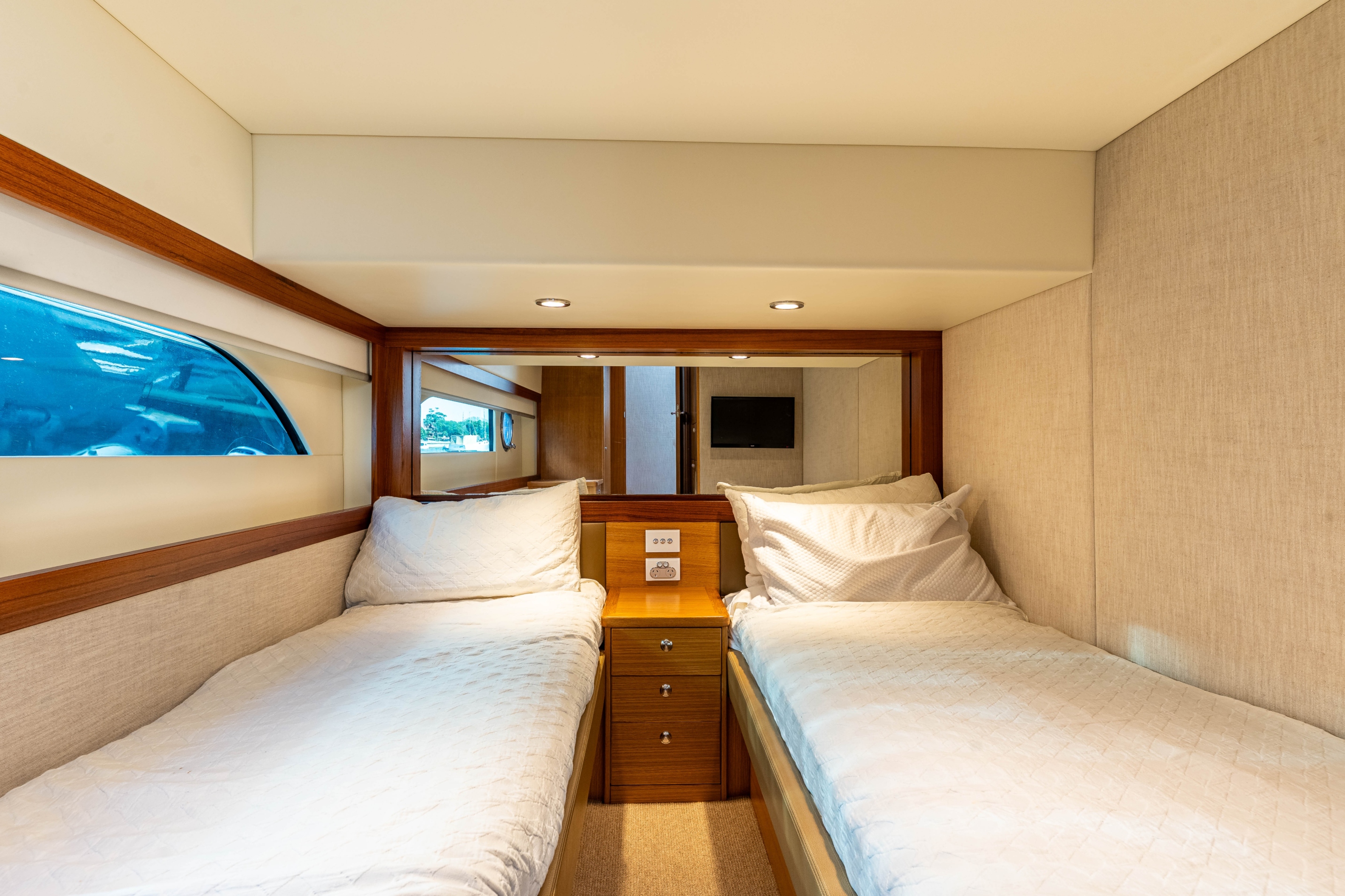 Compact bedroom with two single beds separated by a wooden nightstand, featuring beige bedding and a window with blue tint.