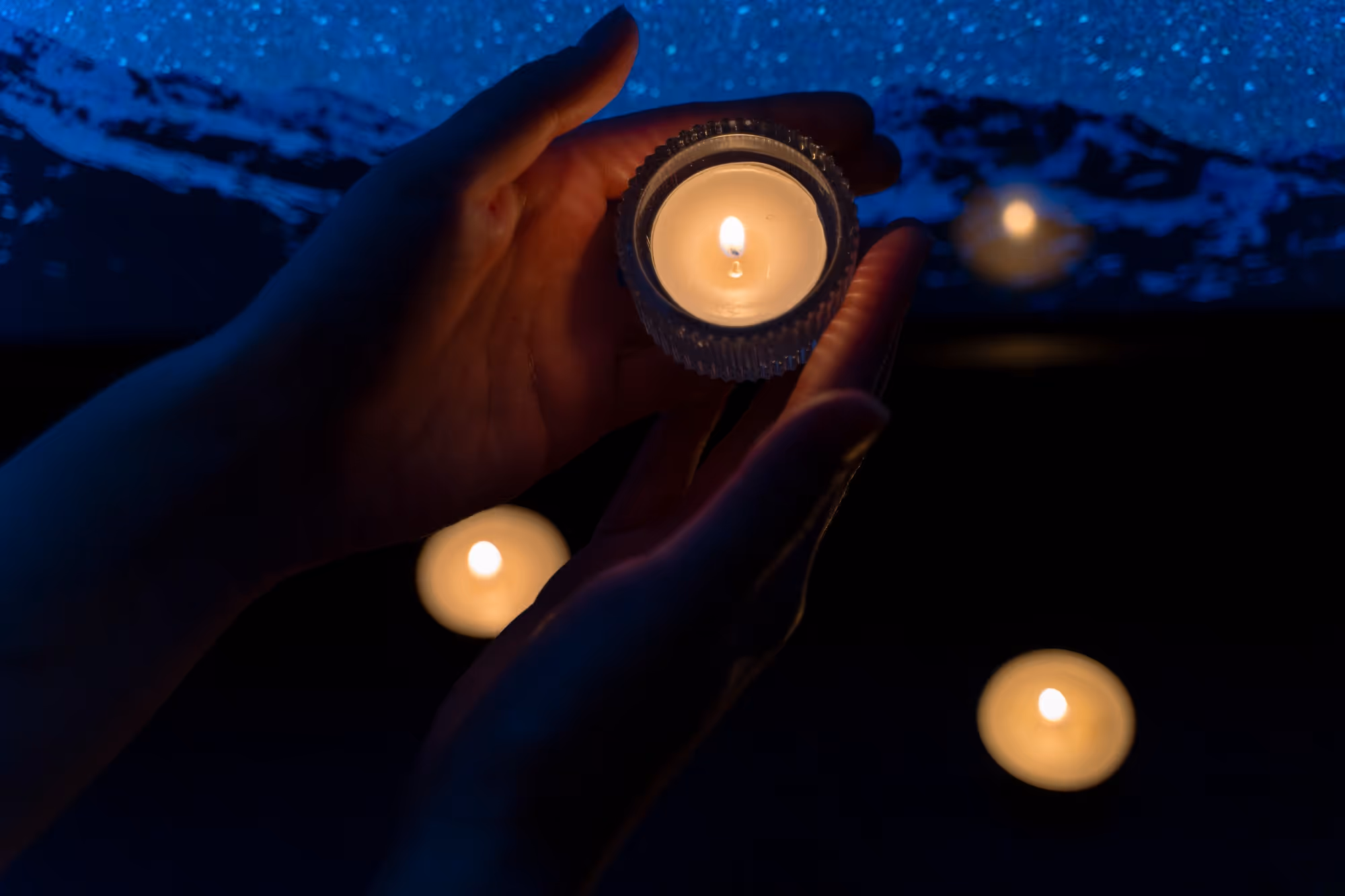 Hands gently holding a lit candle with several other lit candles floating on dark water in the background.