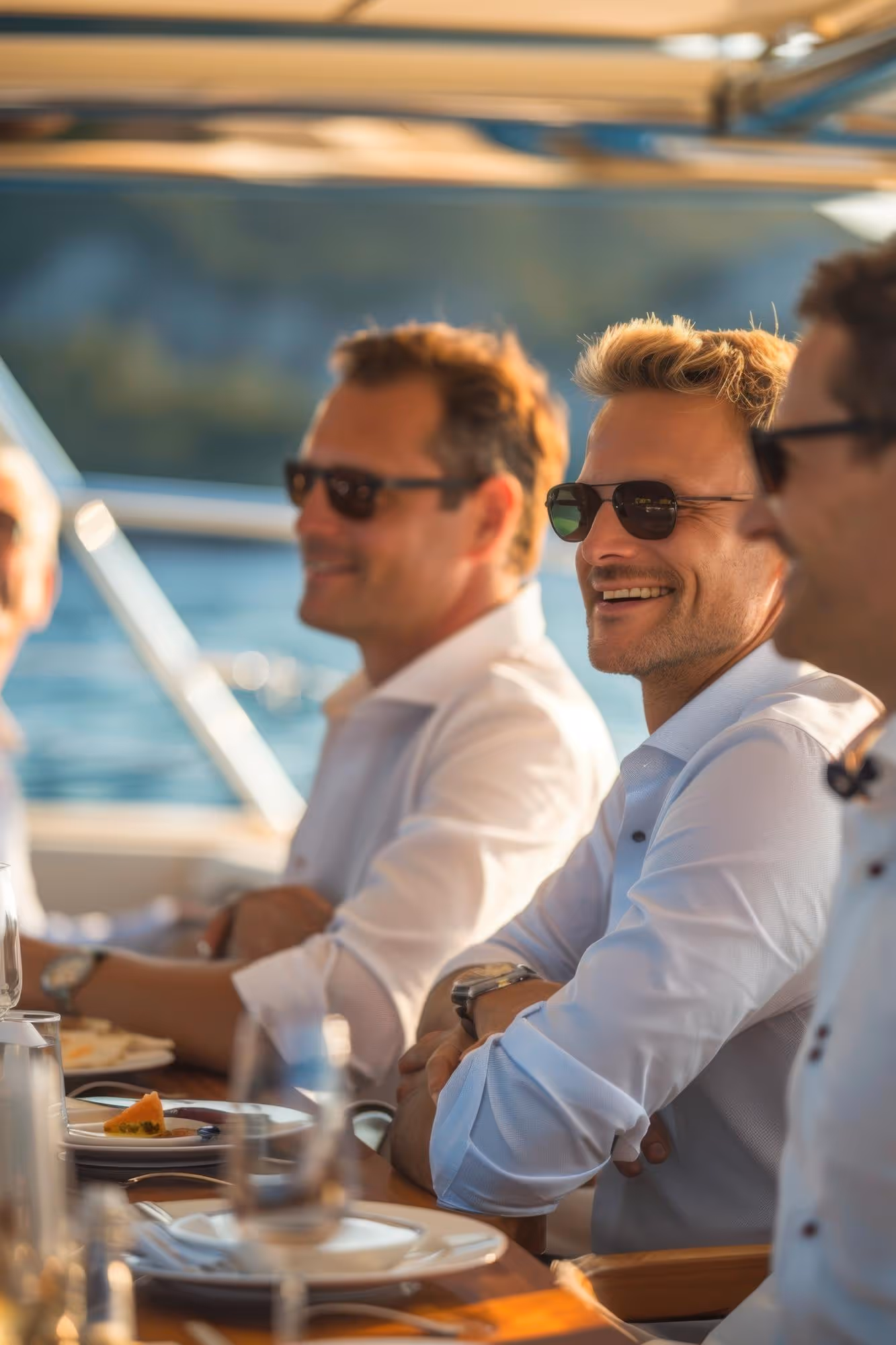 Three men wearing sunglasses and light-colored shirts sitting at a wooden table with plates and glasses, smiling and enjoying a meal outdoors near water.