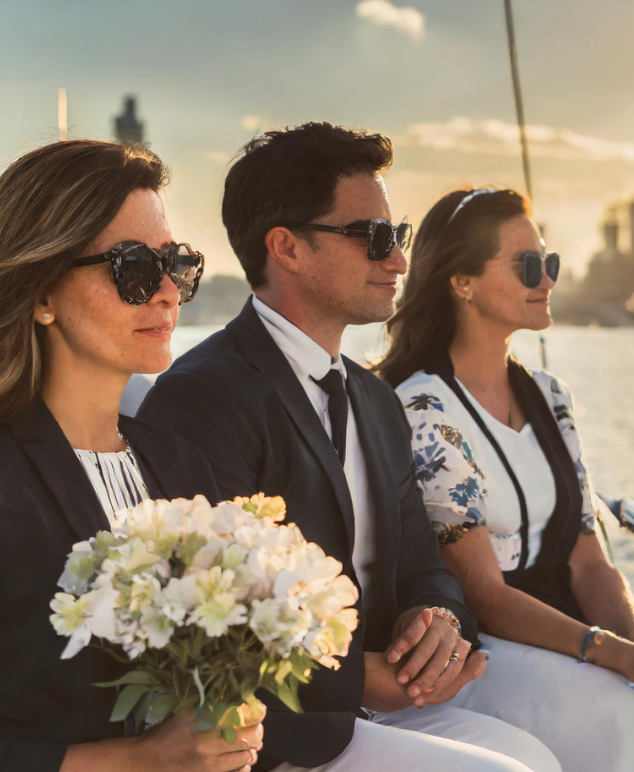 Three people wearing sunglasses sitting closely on a boat at sunset, one holding a bouquet of white flowers.
