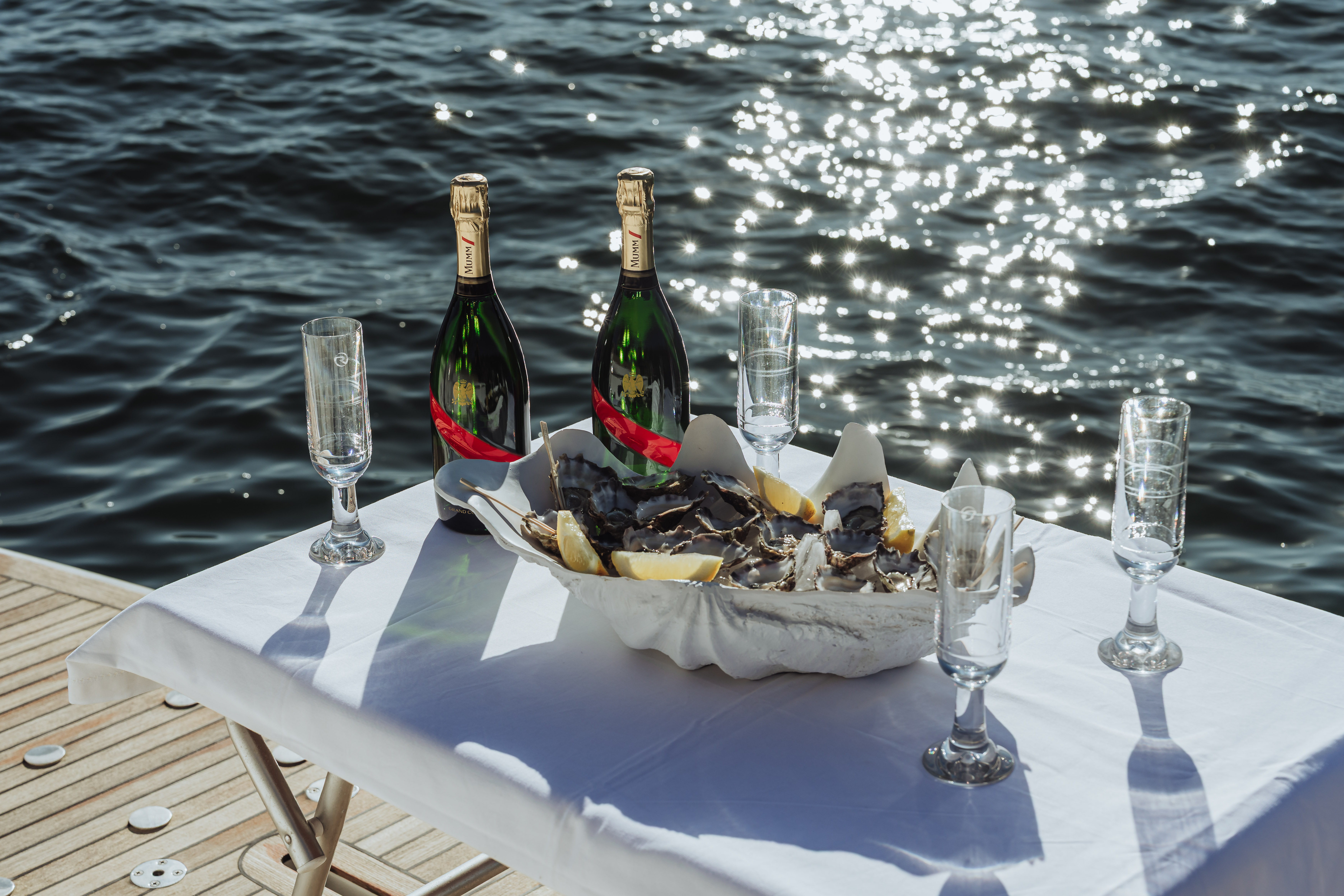 Table with two bottles of champagne, four empty champagne flutes, and a large shell filled with oysters and lemon wedges by sparkling water.