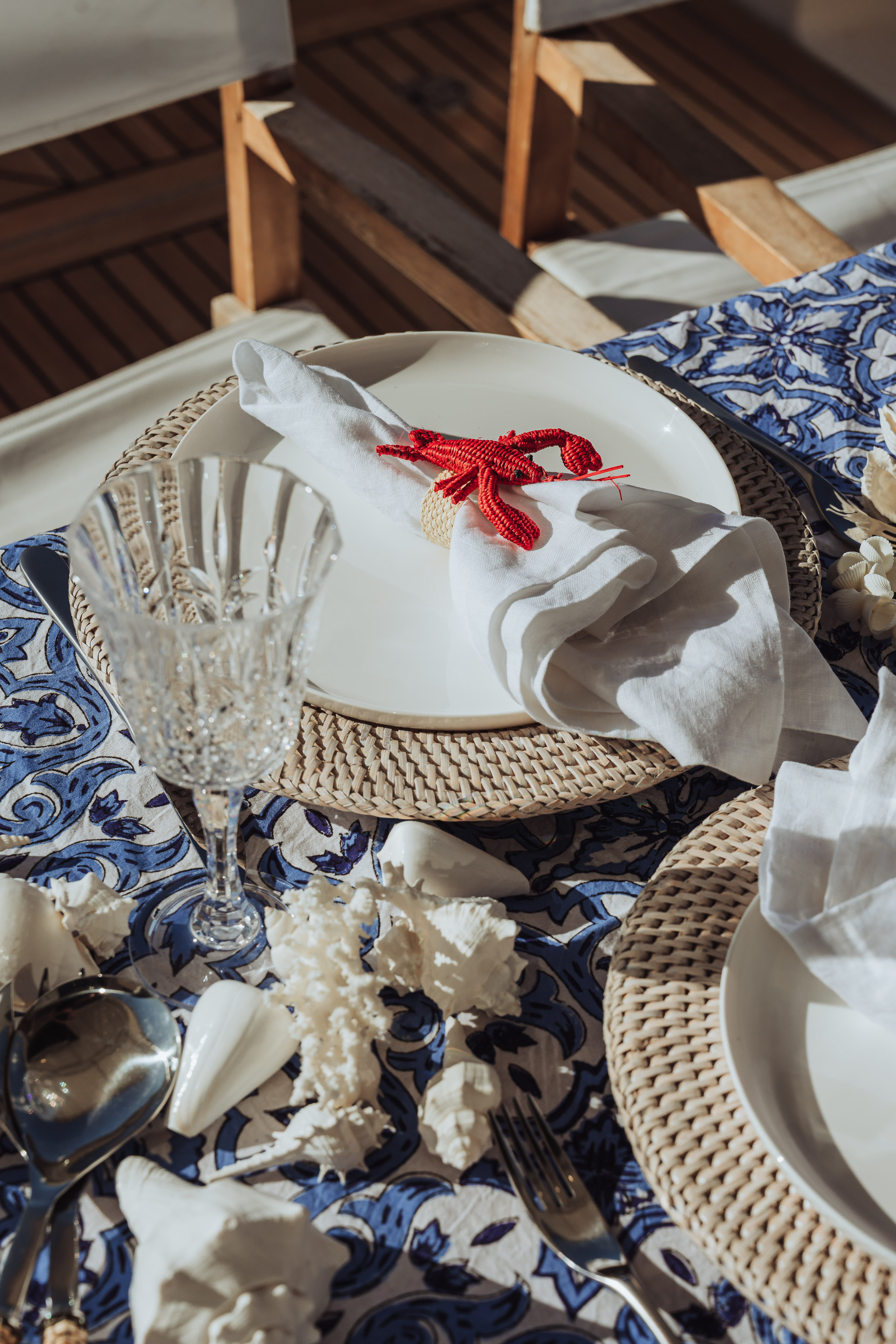 Table set with woven placemats, white plates, crystal wine glass, seashell decorations, and white napkins with a red lobster napkin ring on a blue patterned tablecloth.