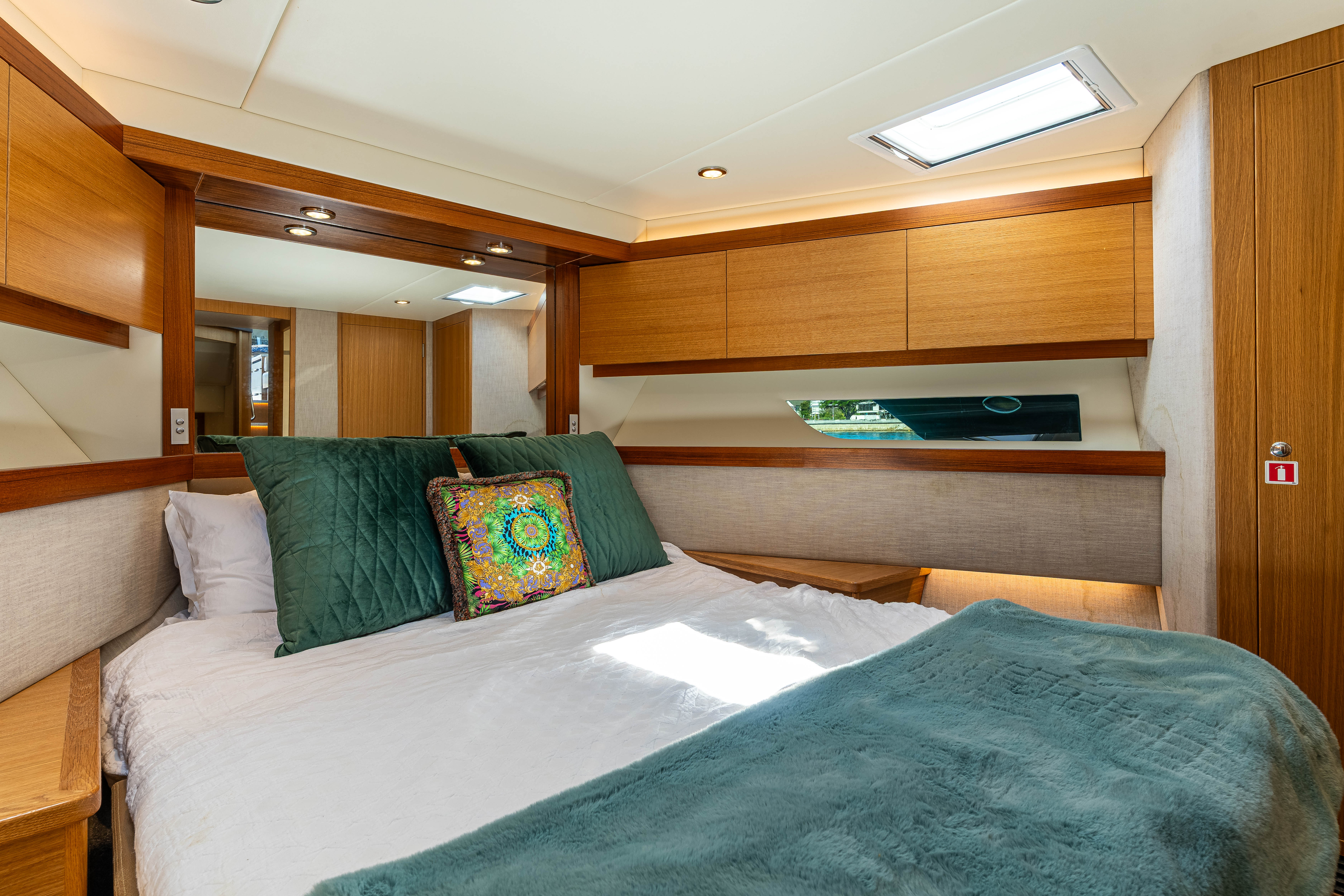 Cozy boat bedroom with white bedding, green pillows, decorative cushion, wood cabinets, and a small window.