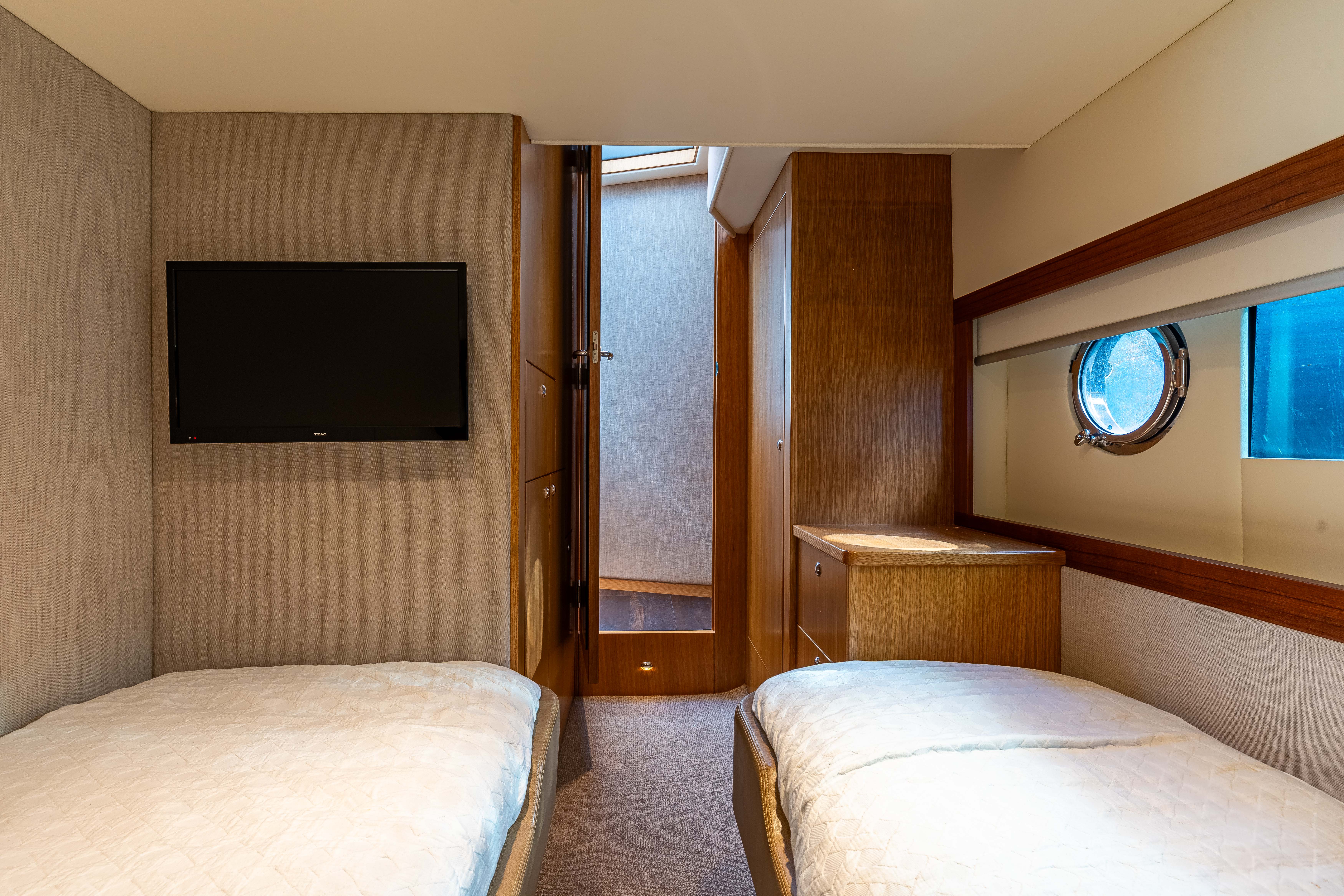 Interior cabin with two single beds, wooden cabinets, a small round window, and a wall-mounted TV.