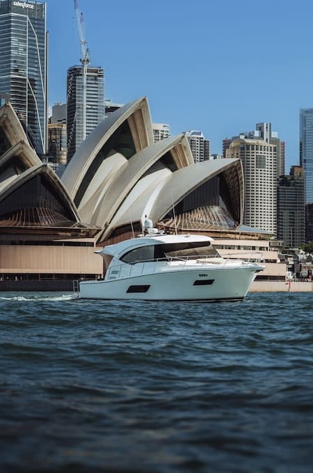 White yacht sailing on water with Sydney Opera House and city skyscrapers in the background under a clear blue sky.