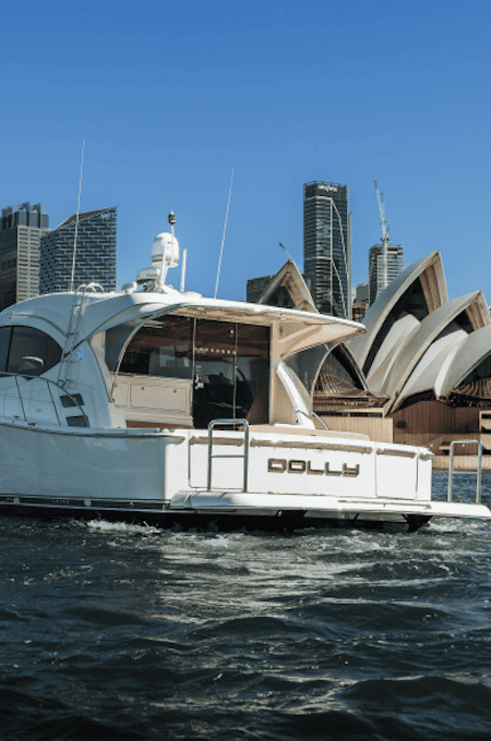 White yacht named 'Dolly' on water with Sydney Opera House and city skyscrapers in the background under clear blue sky.
