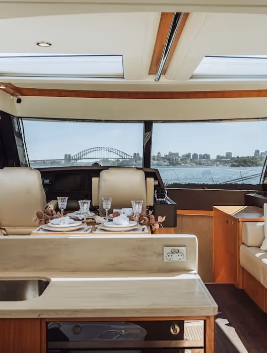 Interior view of a yacht's dining and helm area with table set for four and Sydney Harbour Bridge visible through front windows.