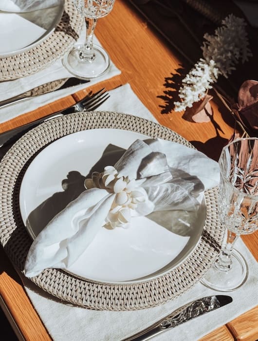 Table setting with white plate on woven placemat, white cloth napkin with decorative rings, crystal wine glass, and silver cutlery on wooden table.