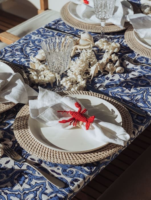Table set with blue patterned tablecloth, woven placemats, white plates, cloth napkins with red lobster napkin rings, and clear crystal glasses.