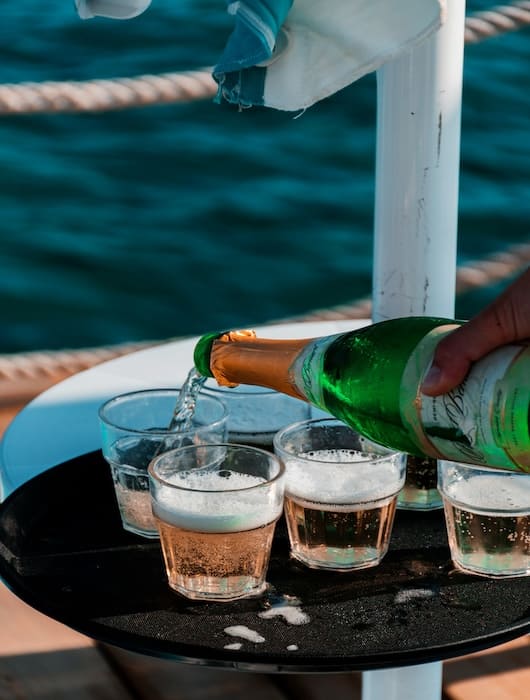 Hand pouring champagne from a green bottle into small glasses on a black tray by the water.