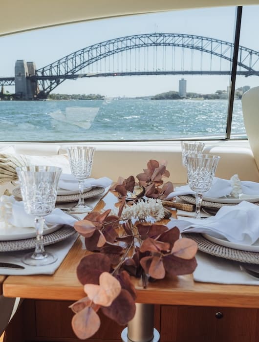 Table set with glassware, white napkins, and dried floral centerpiece on a boat, with Sydney Harbour Bridge visible through the window.