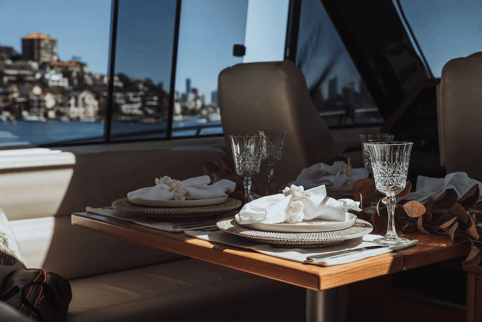 Elegantly set dining table with white napkins and crystal glasses inside a boat, with water and buildings visible through the window.