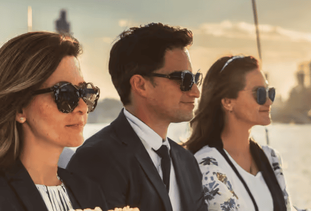 Three people wearing sunglasses and business attire sitting side by side outdoors by the water at sunset.