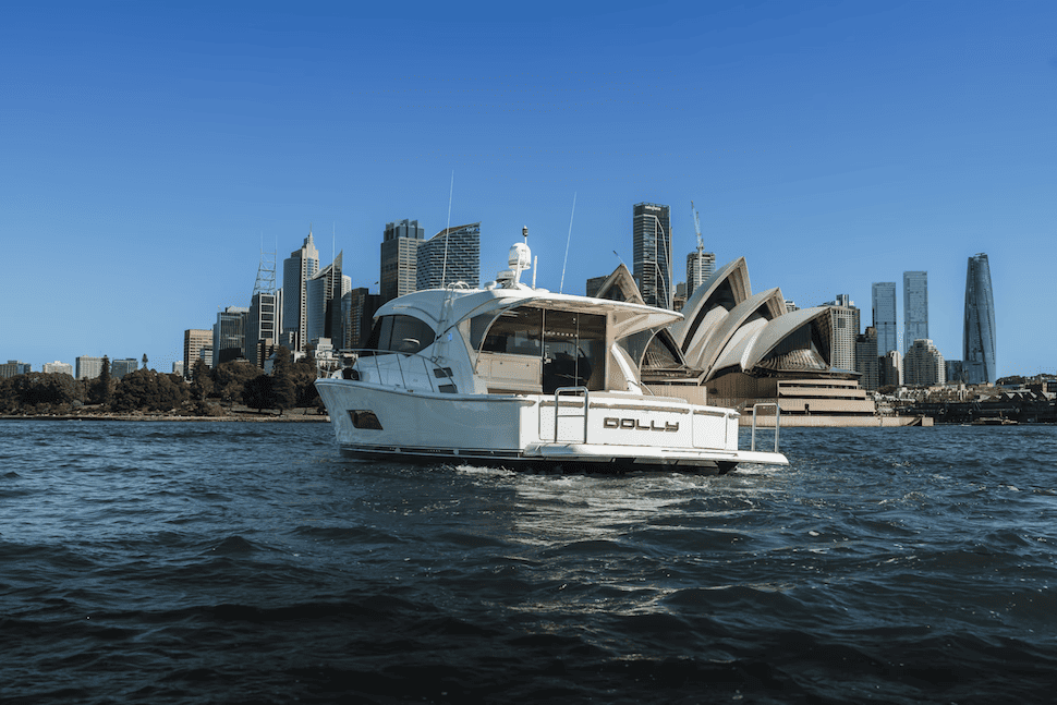 White motor yacht named Dolly sailing on water with Sydney Opera House and city skyline in the background under clear blue sky.