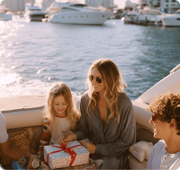 A woman and a young girl smiling and holding a wrapped gift with a red ribbon on a boat, with water and yachts in the background.