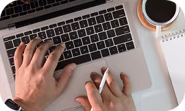 Person typing on laptop keyboard with a stylus in hand, coffee cup and notebook beside.