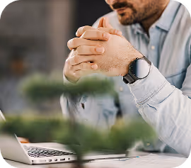 Man wearing a smartwatch and clasping hands in front of a laptop with a blurred indoor background.