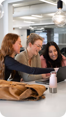 Three women sitting at a table, smiling and looking at a laptop together in a modern office setting.