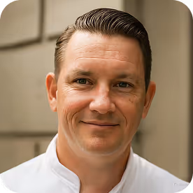 Smiling man with neatly combed dark hair wearing a white collared shirt against a blurred neutral background.