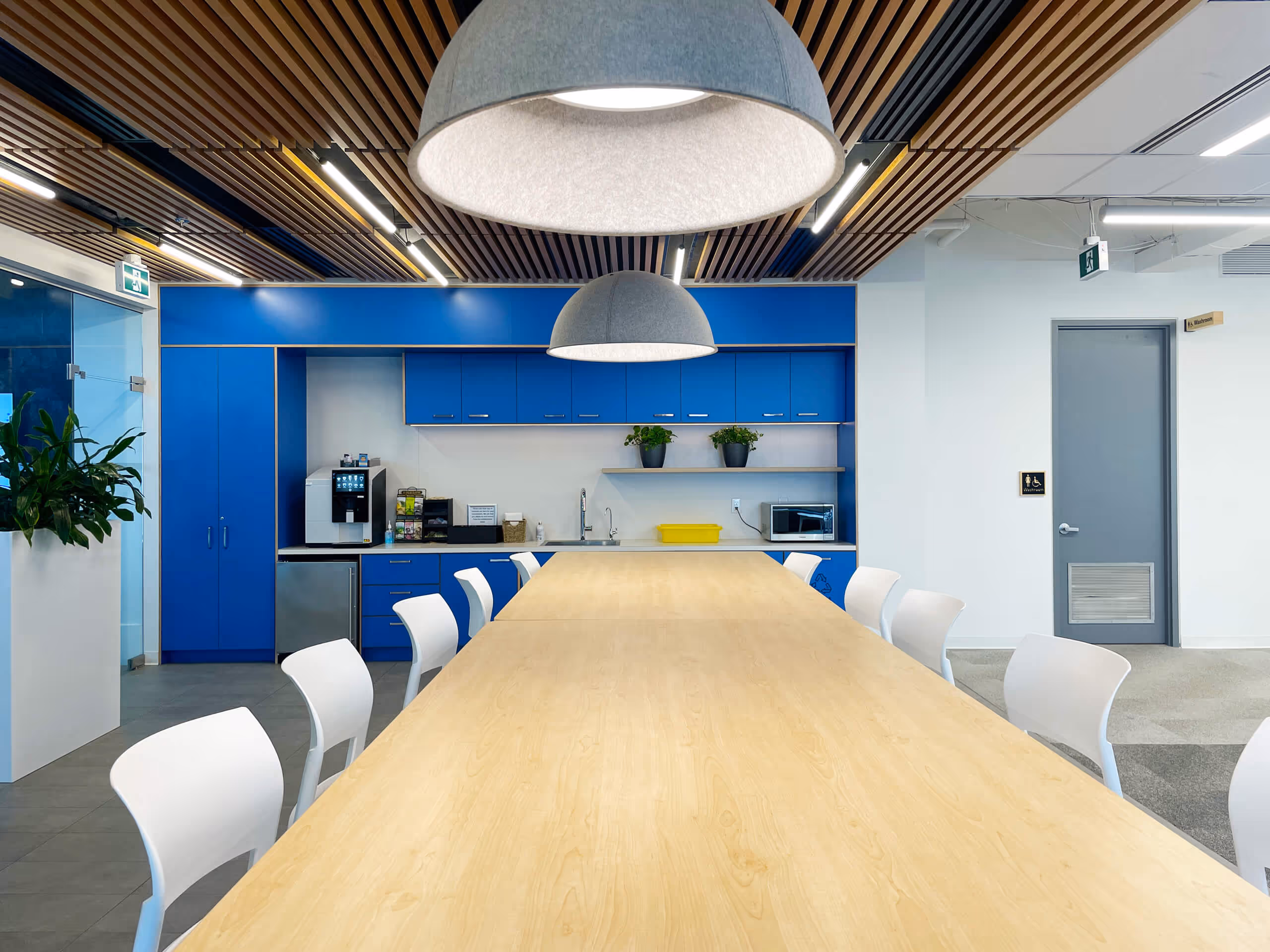 Modern office kitchen with a long wooden table, white chairs, blue cabinets, two hanging gray lamps, and plants on a shelf.
