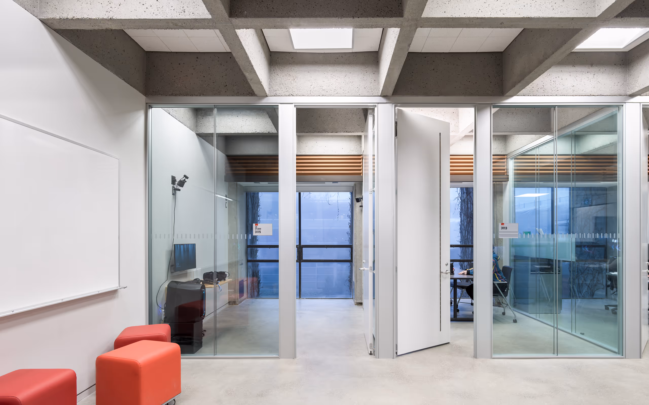 Modern office hallway with glass-walled rooms, concrete ceiling beams, red cube stools, and a whiteboard on the left wall.
