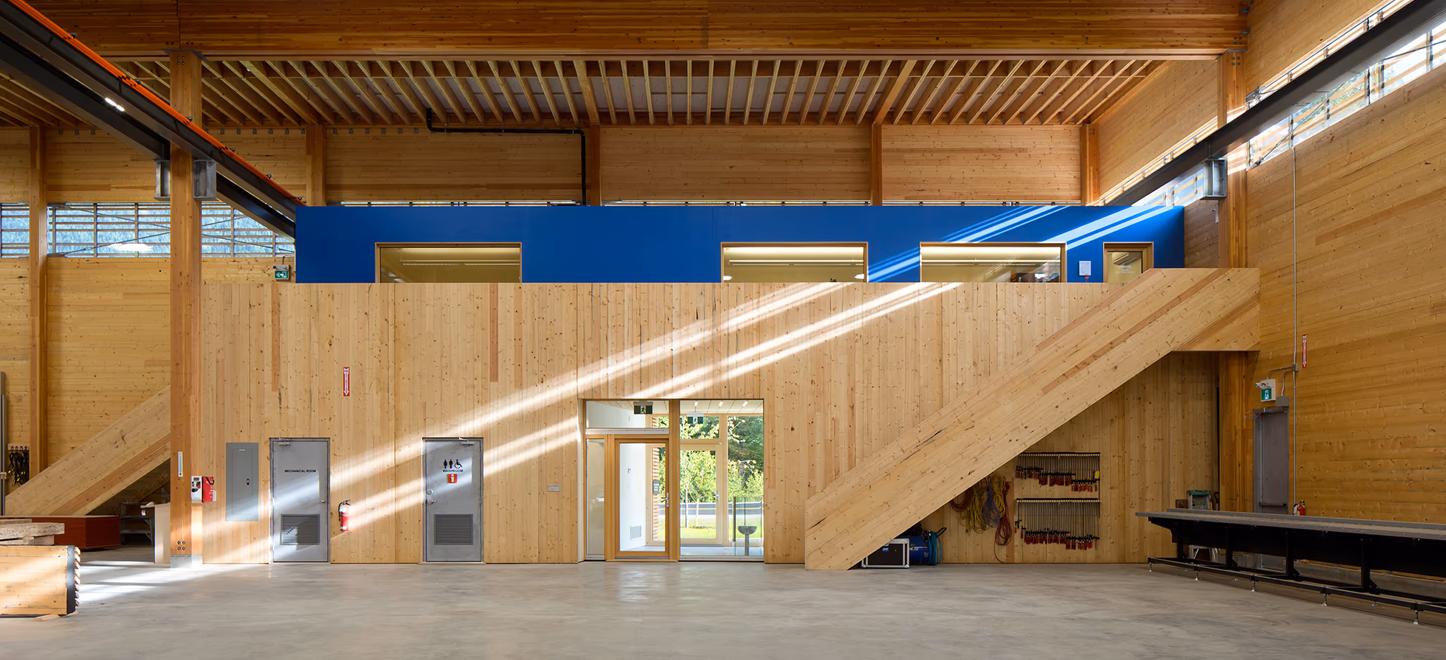 Interior of a spacious wooden building with a large staircase, metal doors labeled mechanical room and restroom, and a blue upper section with windows.