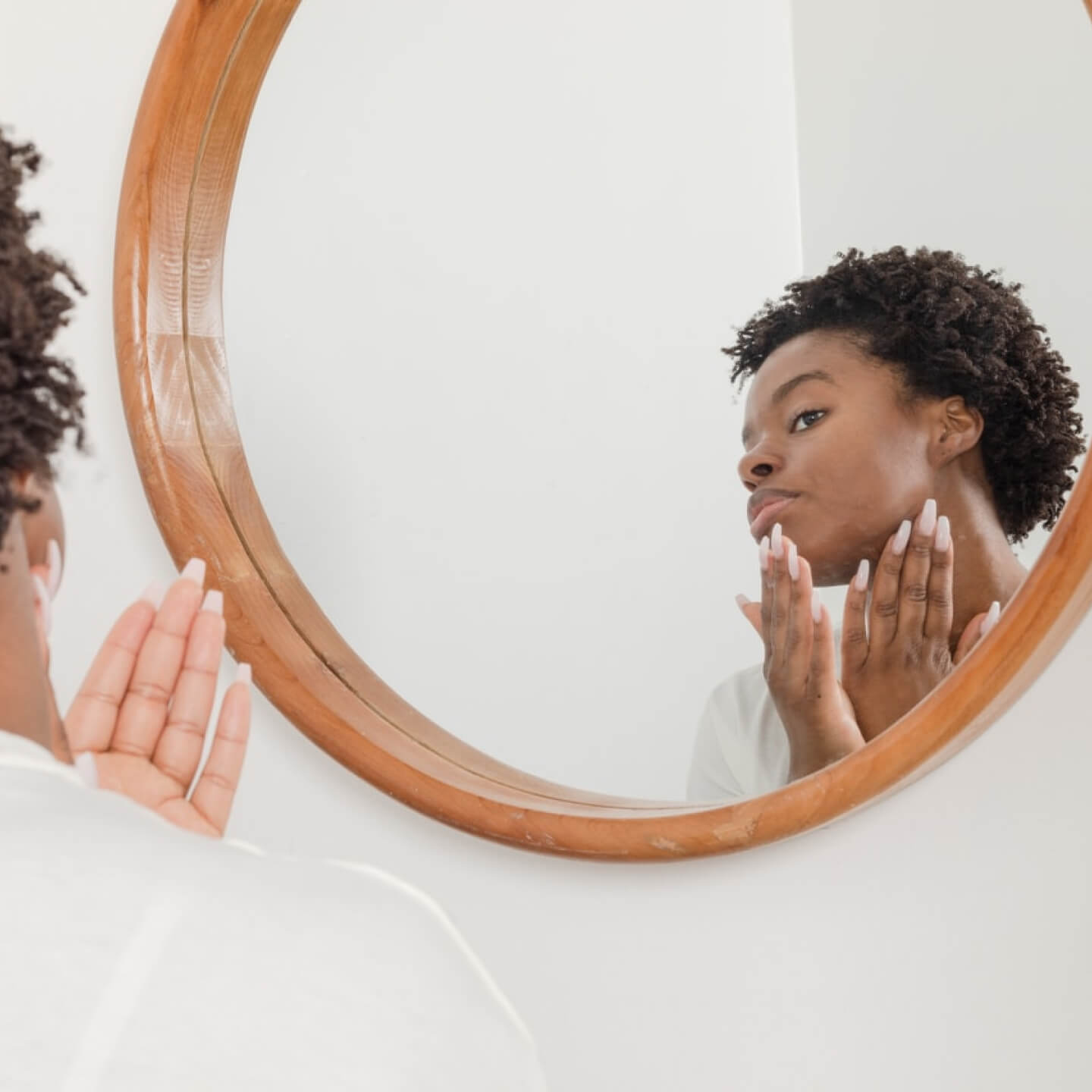 Young woman with curly hair examining her face closely in a round wooden-framed mirror.