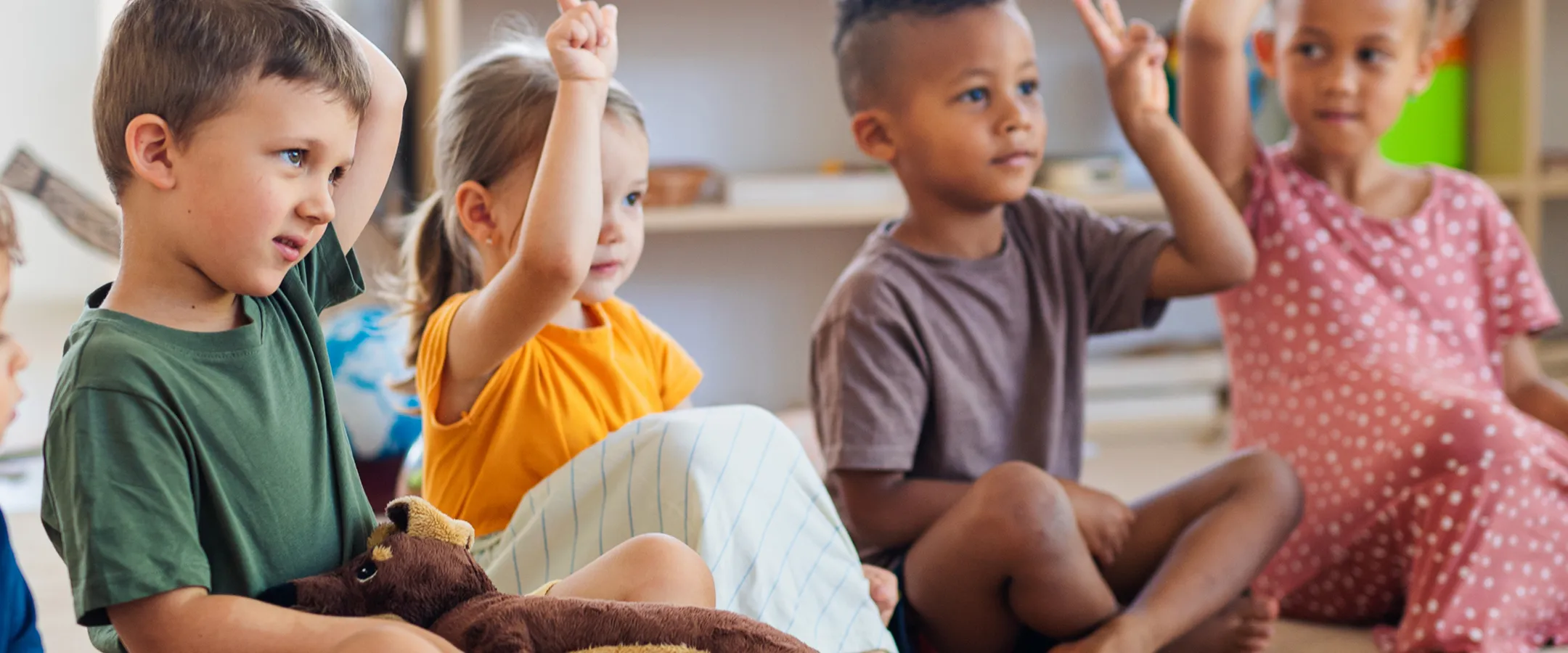 Group of four young children sitting on the floor in a classroom with hands raised.