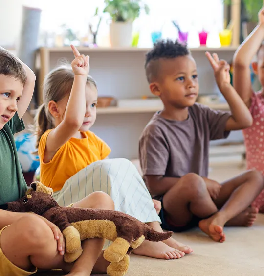 Group of young children sitting on the floor in a classroom raising their hands.