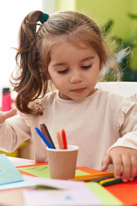 Young girl with a ponytail sitting at a table drawing with colored pencils.