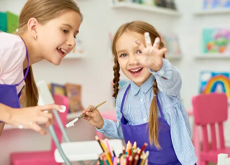 Two girls in aprons smiling and painting together at an art class with colorful pencils and brushes.