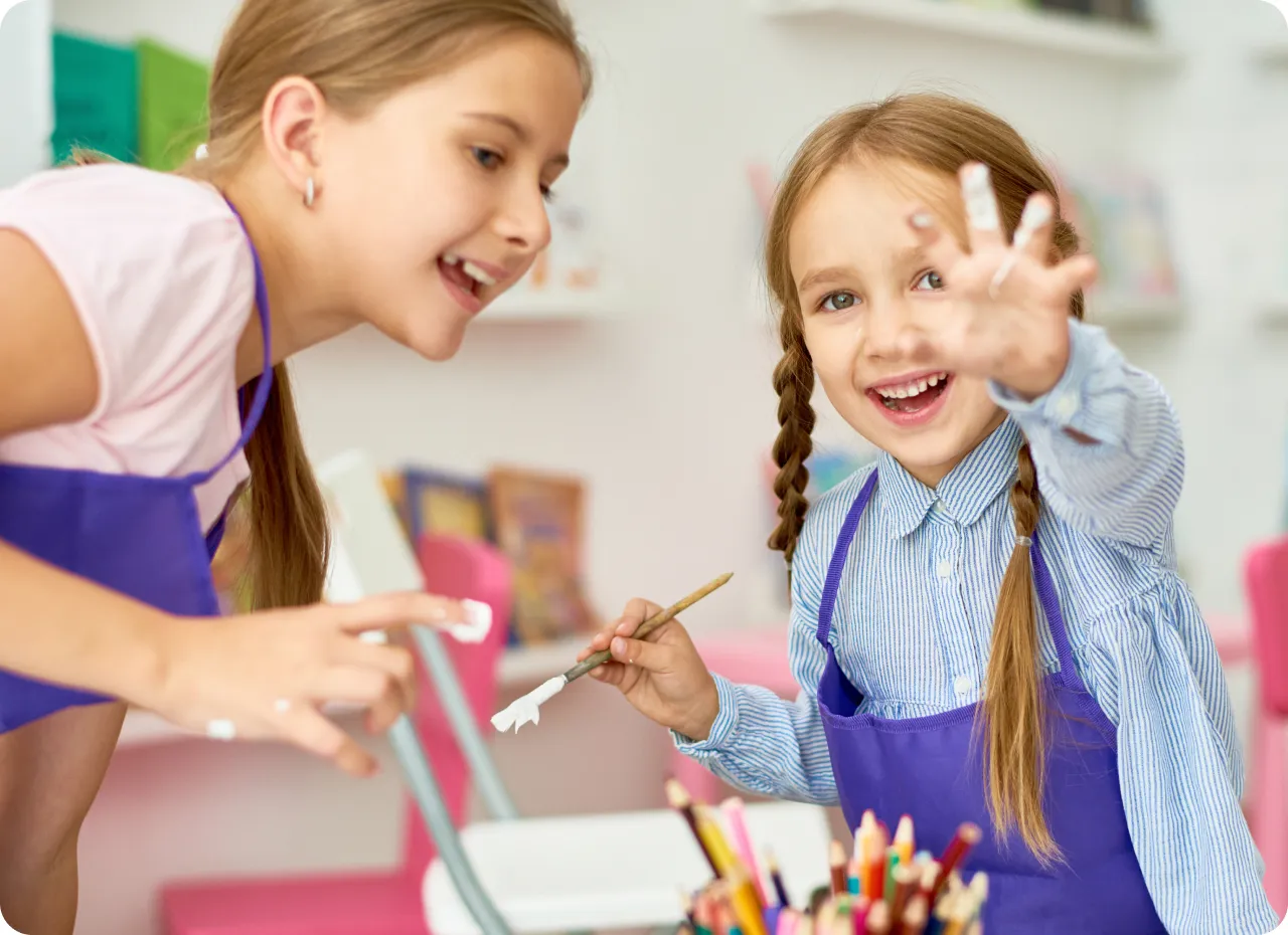 Two smiling young girls wearing purple aprons engaging in painting activities indoors.