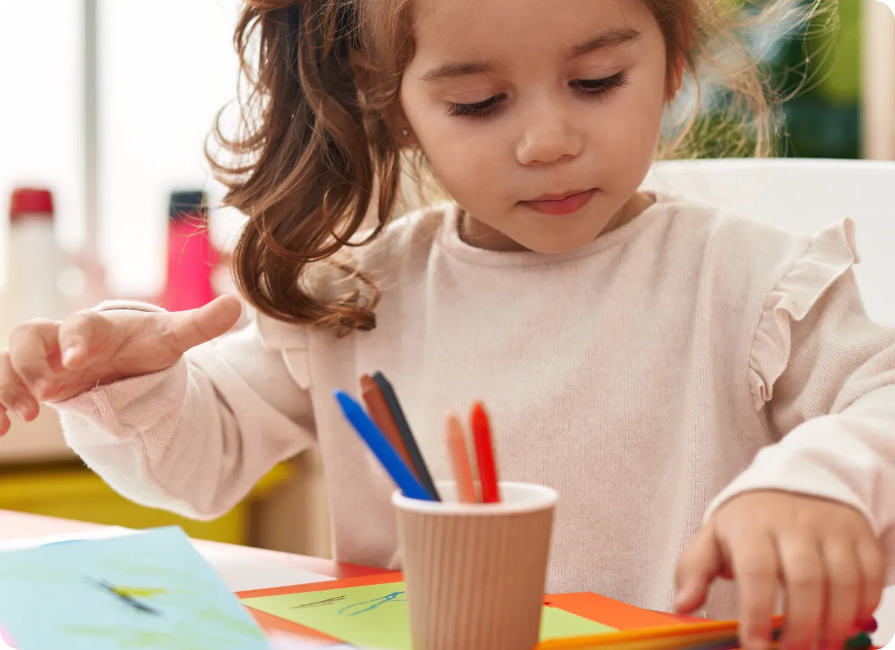 Young girl with brown hair tied in a ponytail focused on drawing at a table with colored paper and markers.