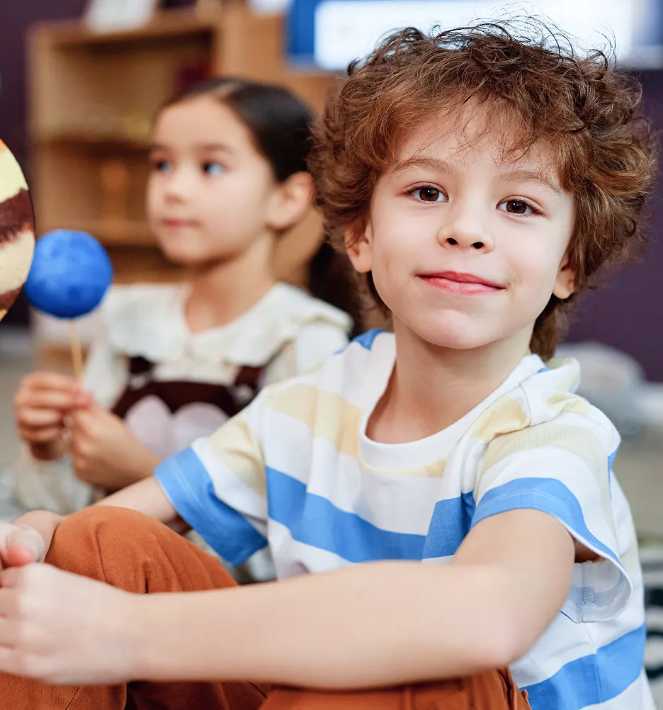 Smiling curly-haired boy in striped shirt sitting in foreground, girl holding planet models blurred in the background.